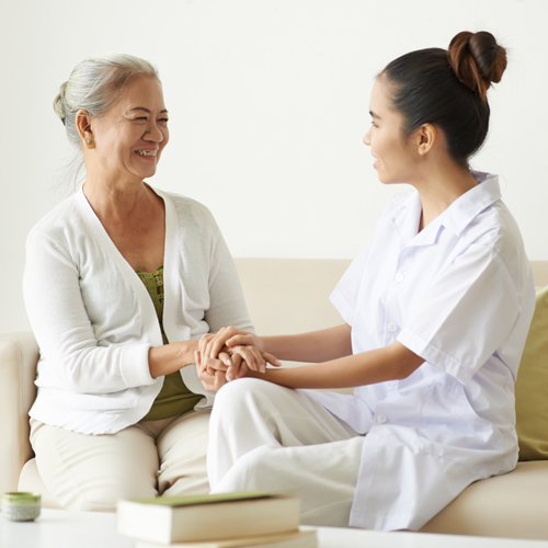 A doctor in a white coat speaking with an older patient in a bright living room
